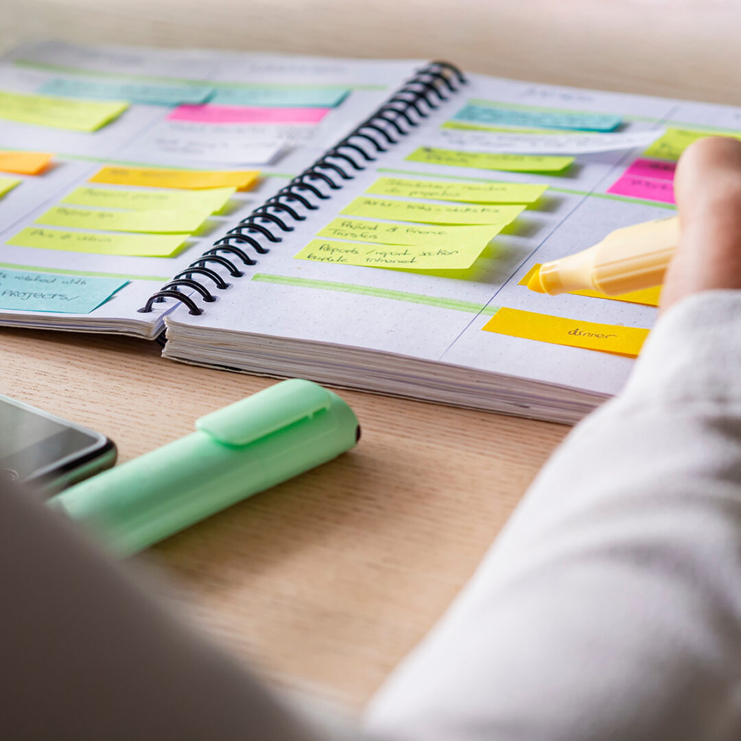 image shows open annual calendar planner with multicolored sticky notes and markers, and one hand writing.