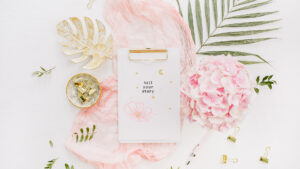 image shows a flatlay with leaves, a pink swath of fabric, gold clips, and a porcelain gold bowl. A clipboard with gold and pink flowers and moons and stars sits on top of the pile of pink and gold things. On the paper are the words, "tell your story"