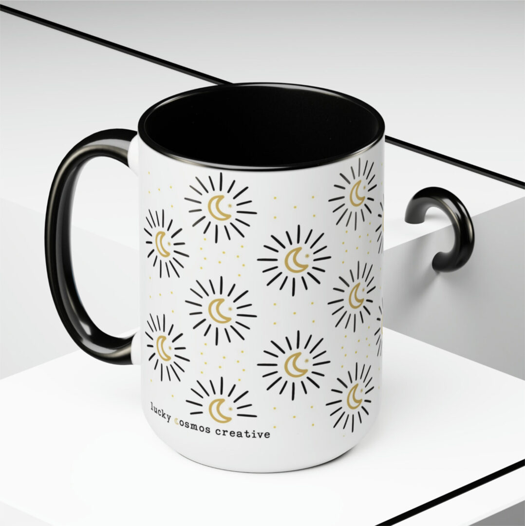 image shows a white mug with a black handle and black inside. The pattern on the mug is gold moons with black sparkles around them. the mug says www.luckycosmoscreative.com