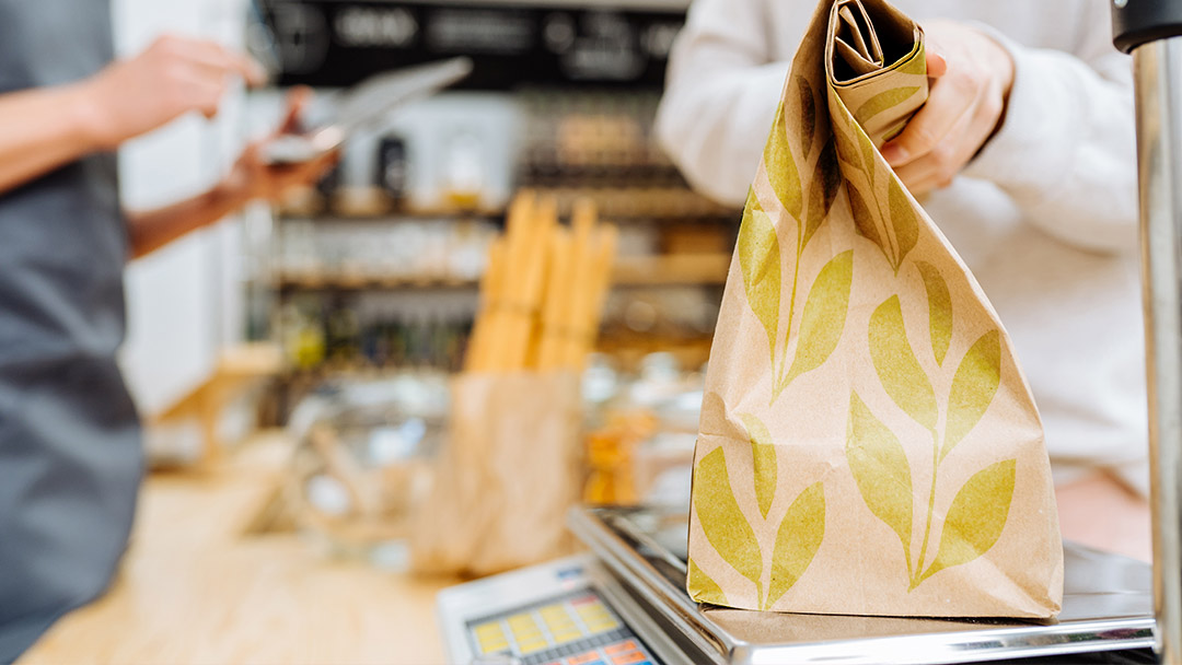Image shows a brown paper shopping bag with hands folding it closed on a store counter, as a visual representation of voting with our dollars.